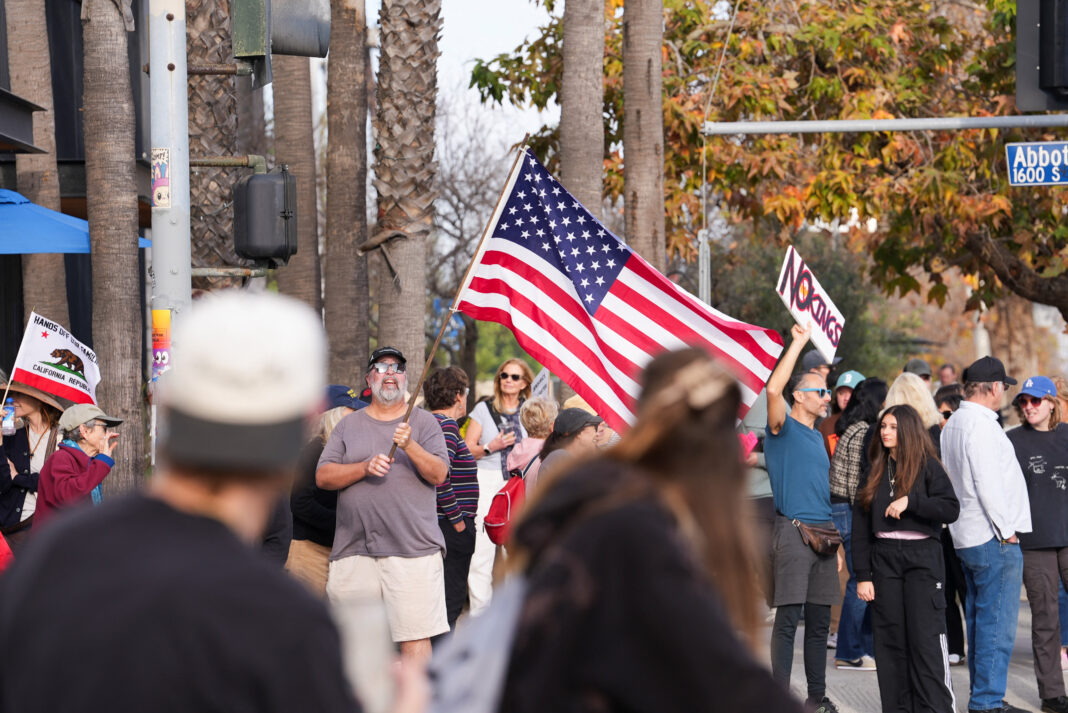 Oamenii țin pancarte și un steag american în timpul unui protest împotriva politicilor președintelui american Donald Trump, la un an de la începutul celui de-al doilea mandat al său, în Los Angeles, California, SUA, 20 ianuarie 2026